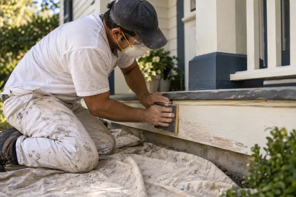 Illustrative exterior preparation scene with a painter sanding porch trim